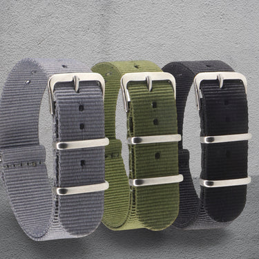 Three NATO watch straps in grey, green, and black with silver buckles on a grey background